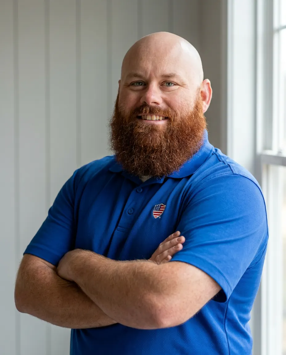 Timothy Ray, founder and owner of Patriot Construction. Bald with a full reddish beard, royal blue Patriot Construction polo, arms crossed, standing inside a residential job site with soft natural daylight from a window on the right
